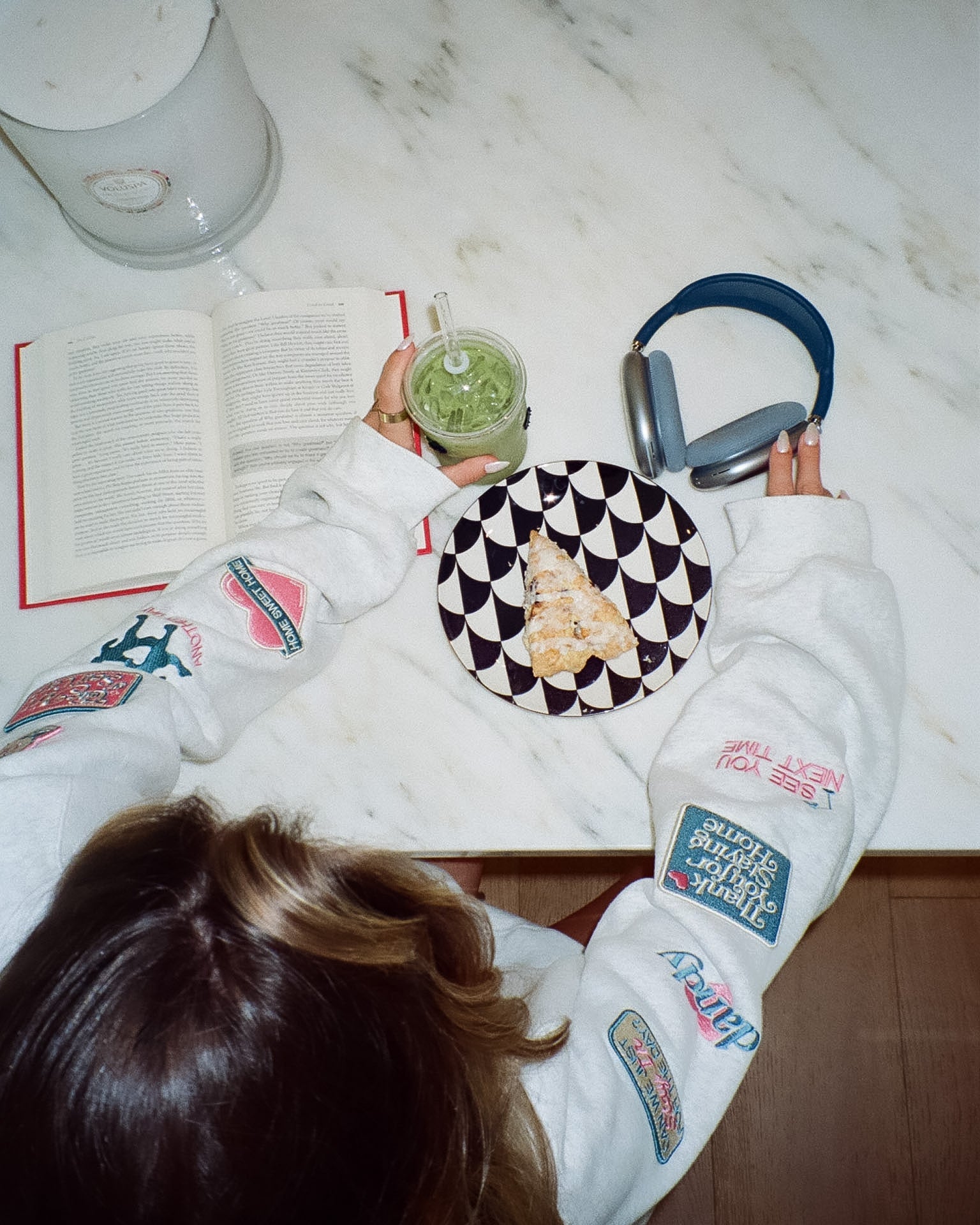 Person sitting at a table with a book, green smoothie, and snacks, wearing a white sweatshirt with colorful patches.