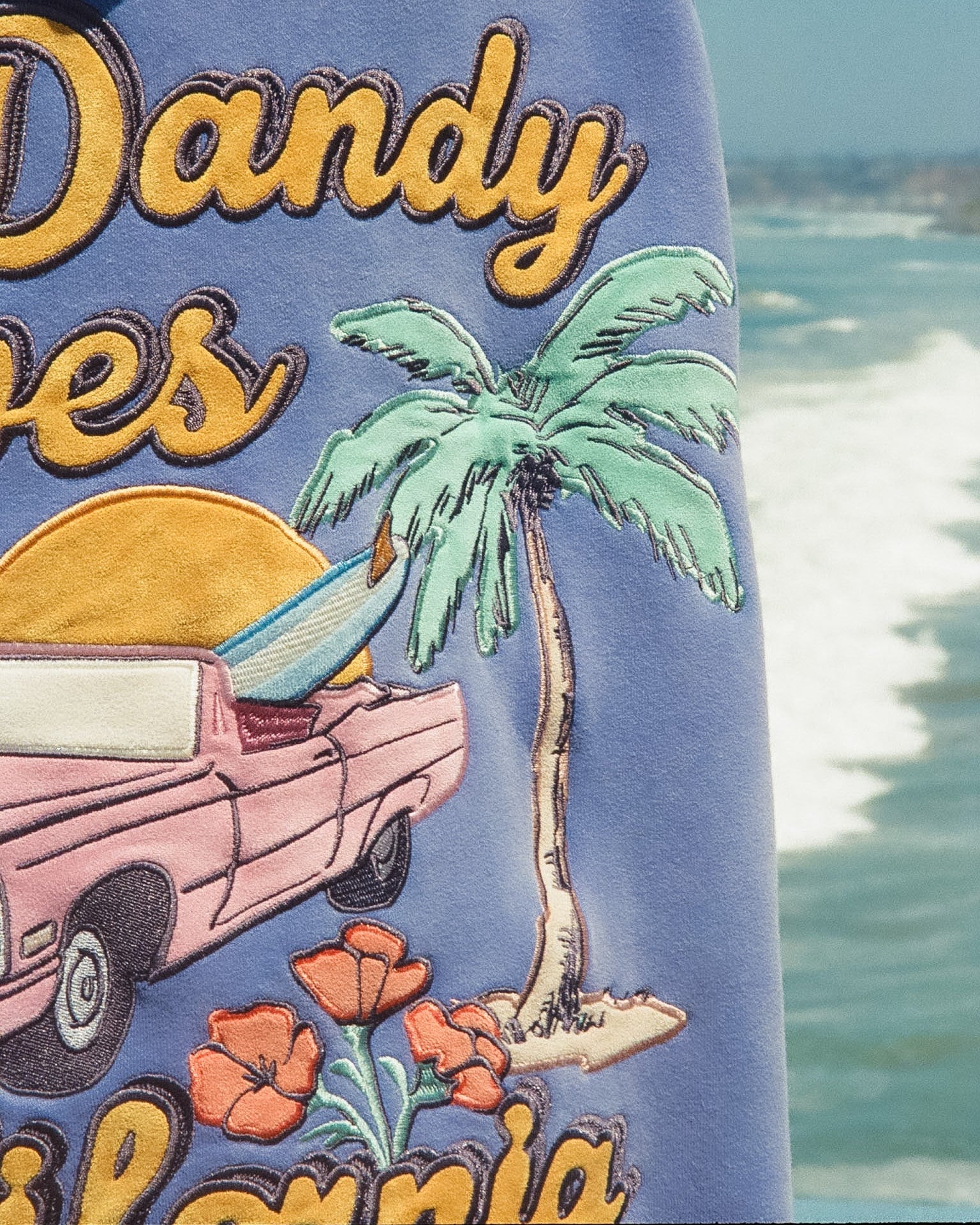 Embroidered design on a fabric with 'Dandy' text, palm tree, and vintage car, against a beach background.