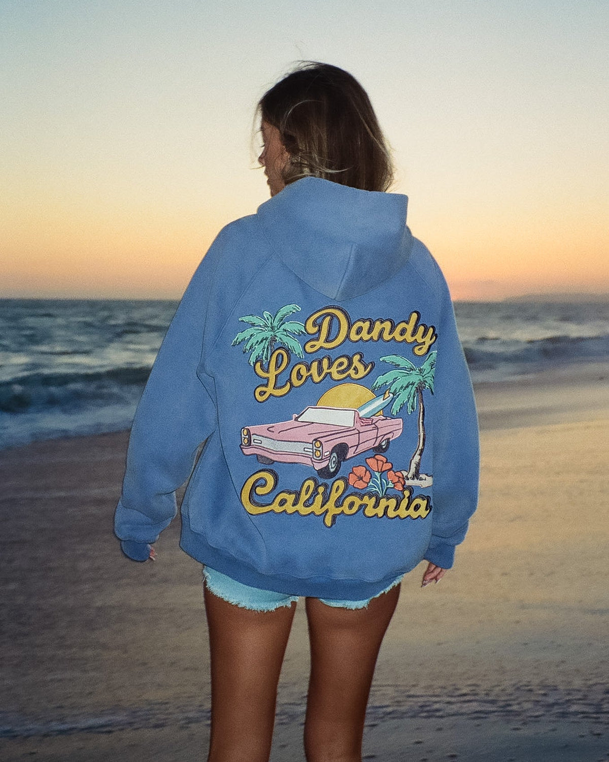 Person wearing a blue hoodie with 'Dandy Loves California' design on a beach at sunset.