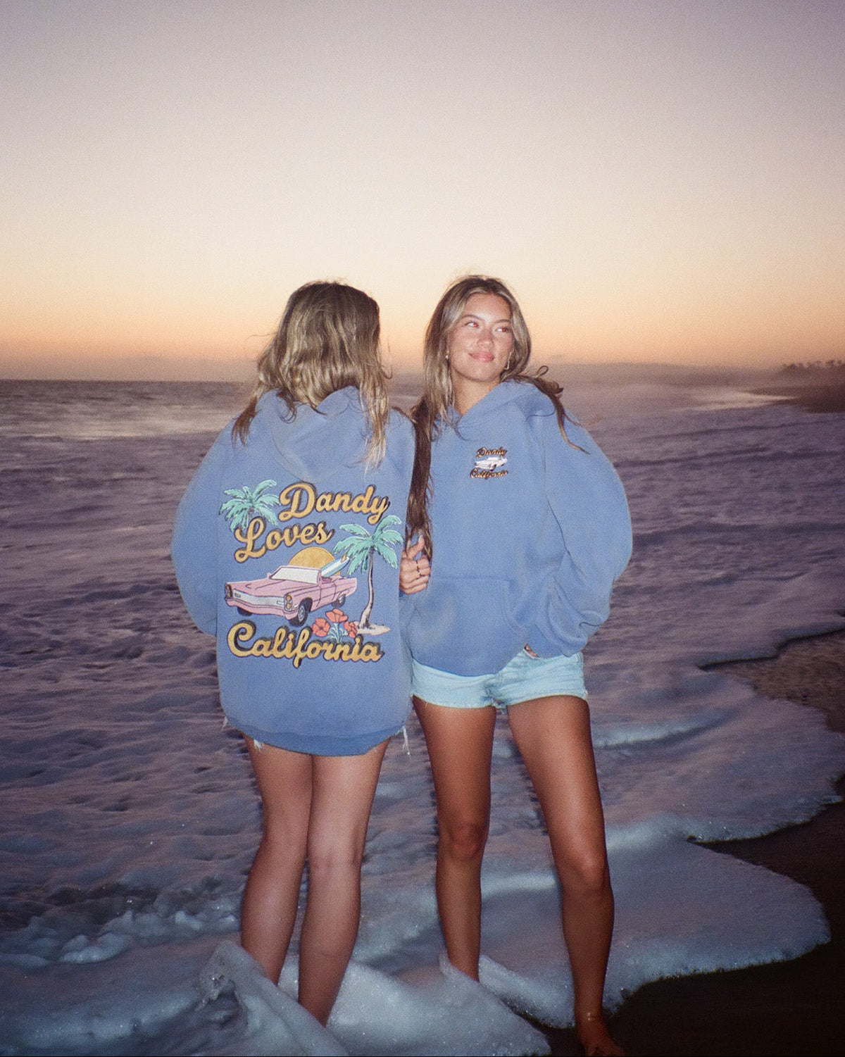 Two people wearing blue hoodies with text and graphics, standing on a beach at sunset.