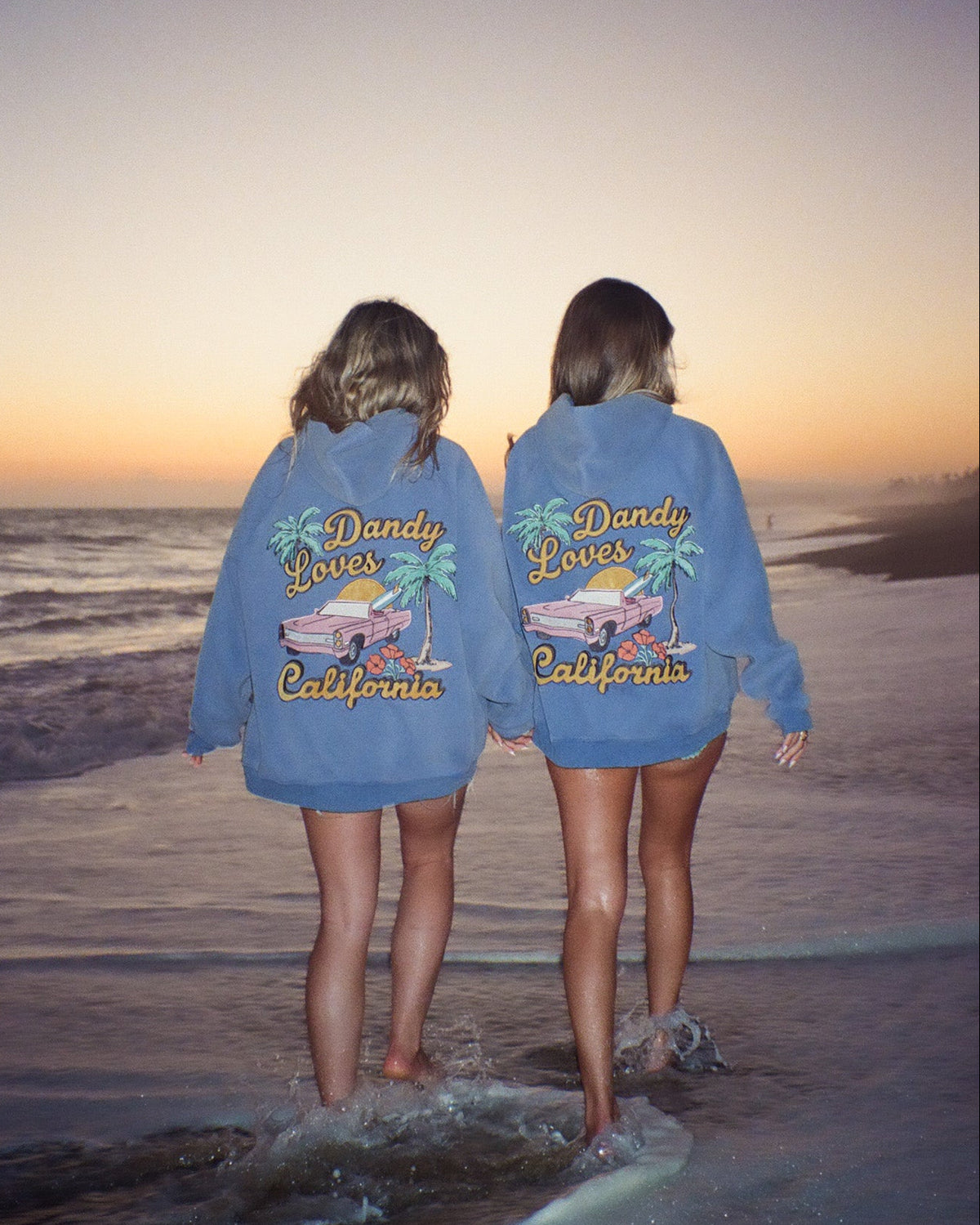 Two people wearing blue sweatshirts with 'Dandy Loves California' text on a beach at sunset.