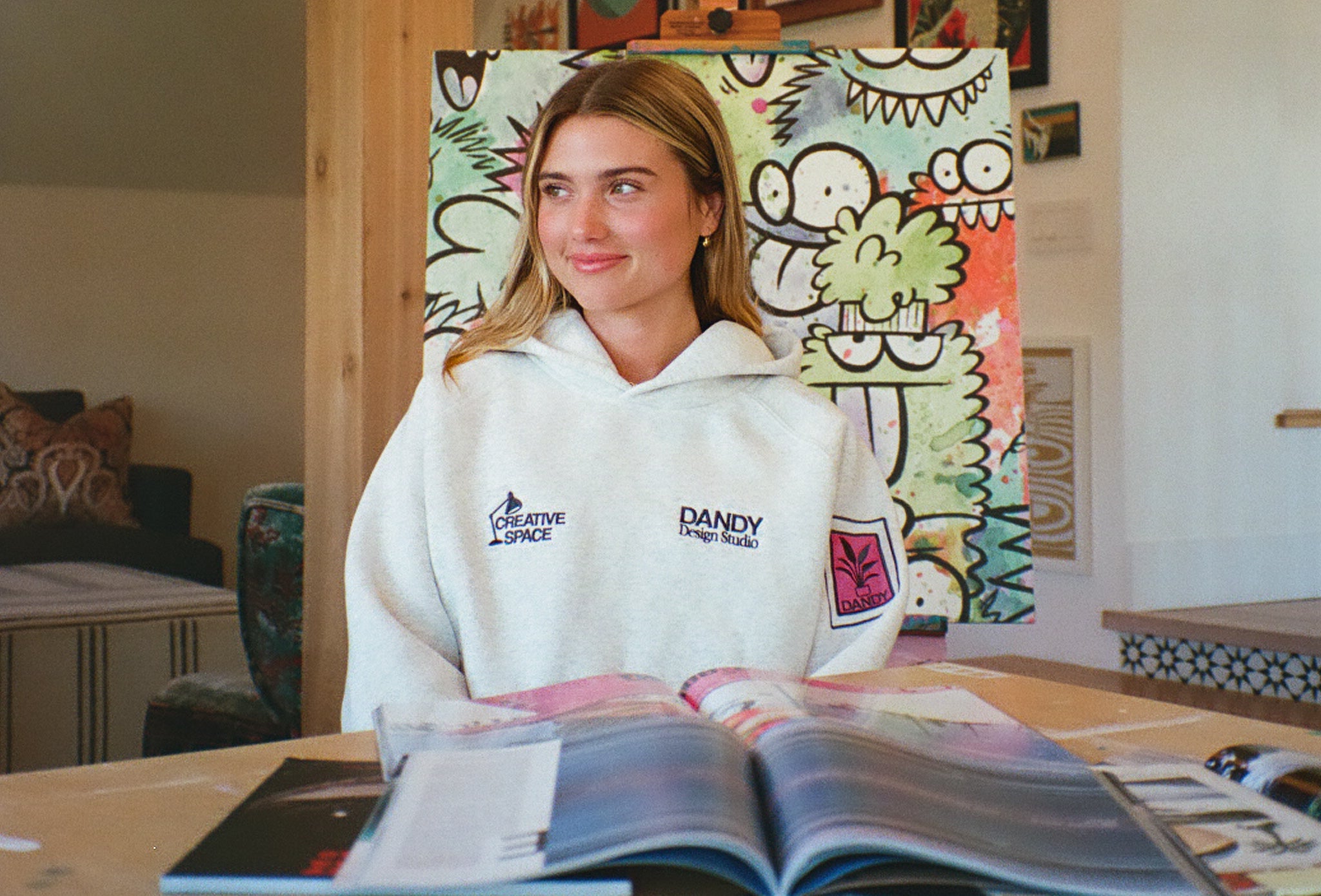 Person wearing a heather grey hoodie with DANDY logo, sitting at a table with an open book in a room with colorful wall art.