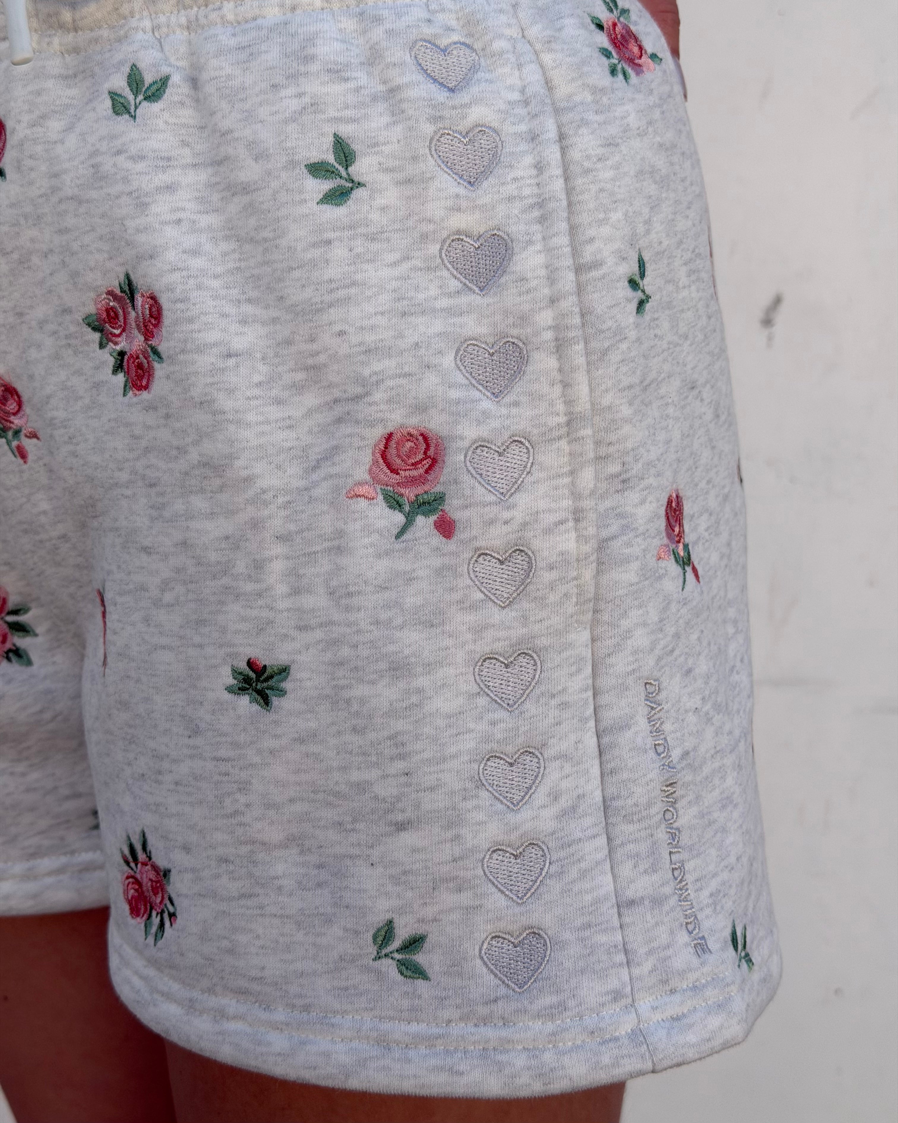 Close-up of light heather gray shorts with embroidered pink roses and green leaves, plus a vertical row of outlined hearts down the front side.