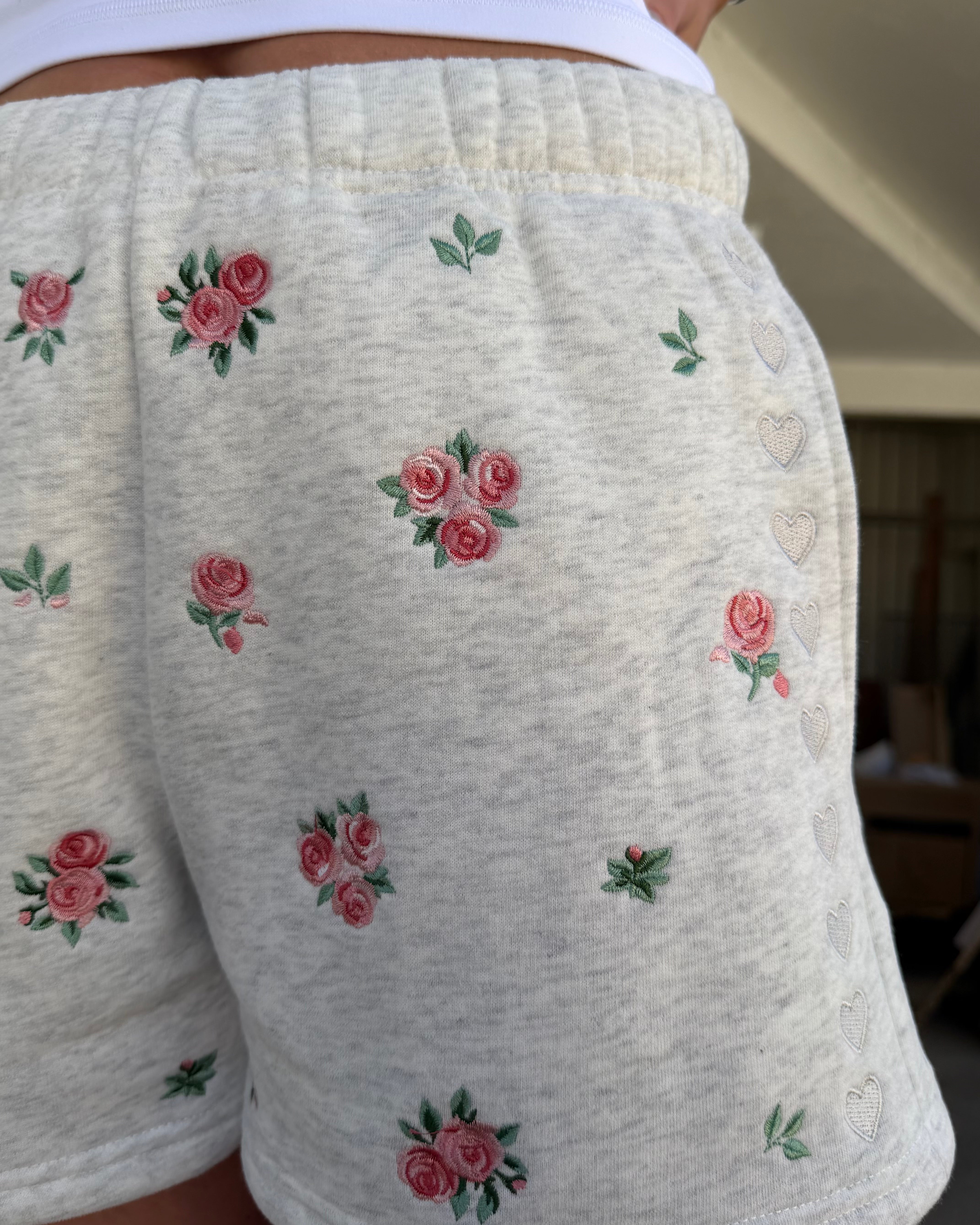 Close-up of a person wearing light heather gray shorts with embroidered pink rose clusters and small heart motifs.