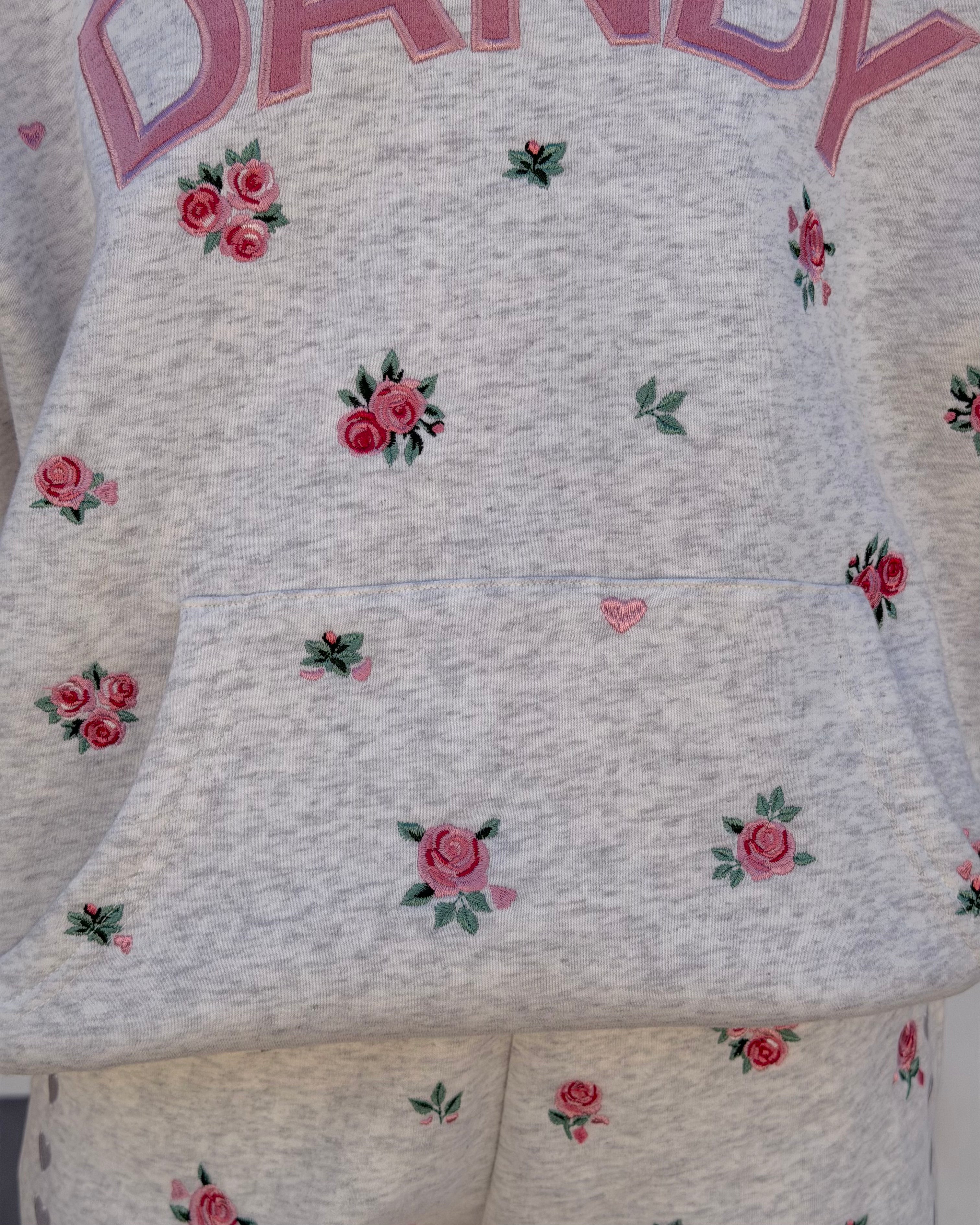 Close-up of a light heather gray sweatshirt with pink embroidered roses and green leaves, featuring a front pocket and partial pink lettering at the top.