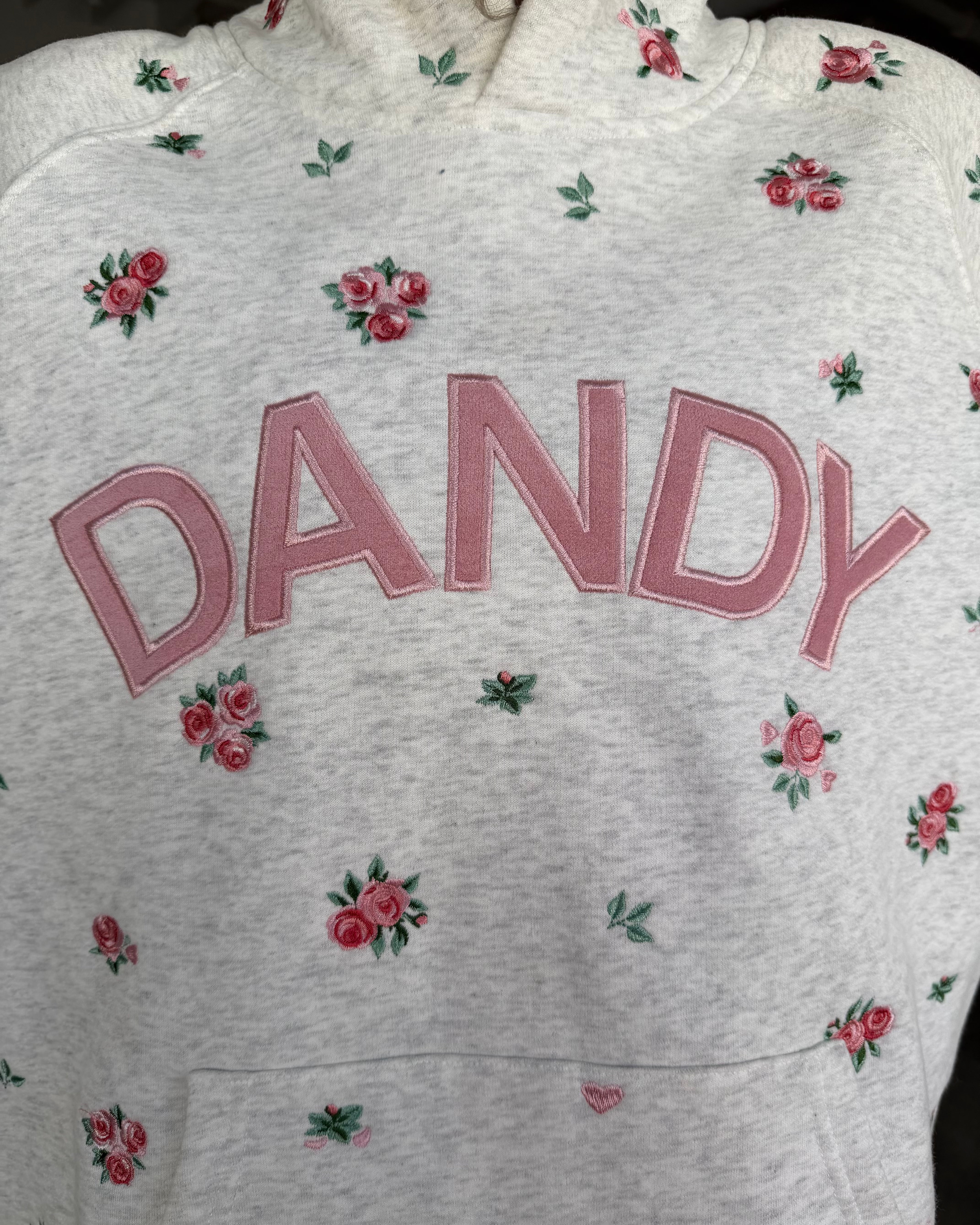 A close-up of a light heather gray sweatshirt featuring the word 'DANDY' embroidered in pink across the chest, surrounded by small pink rose and green leaf floral motifs.