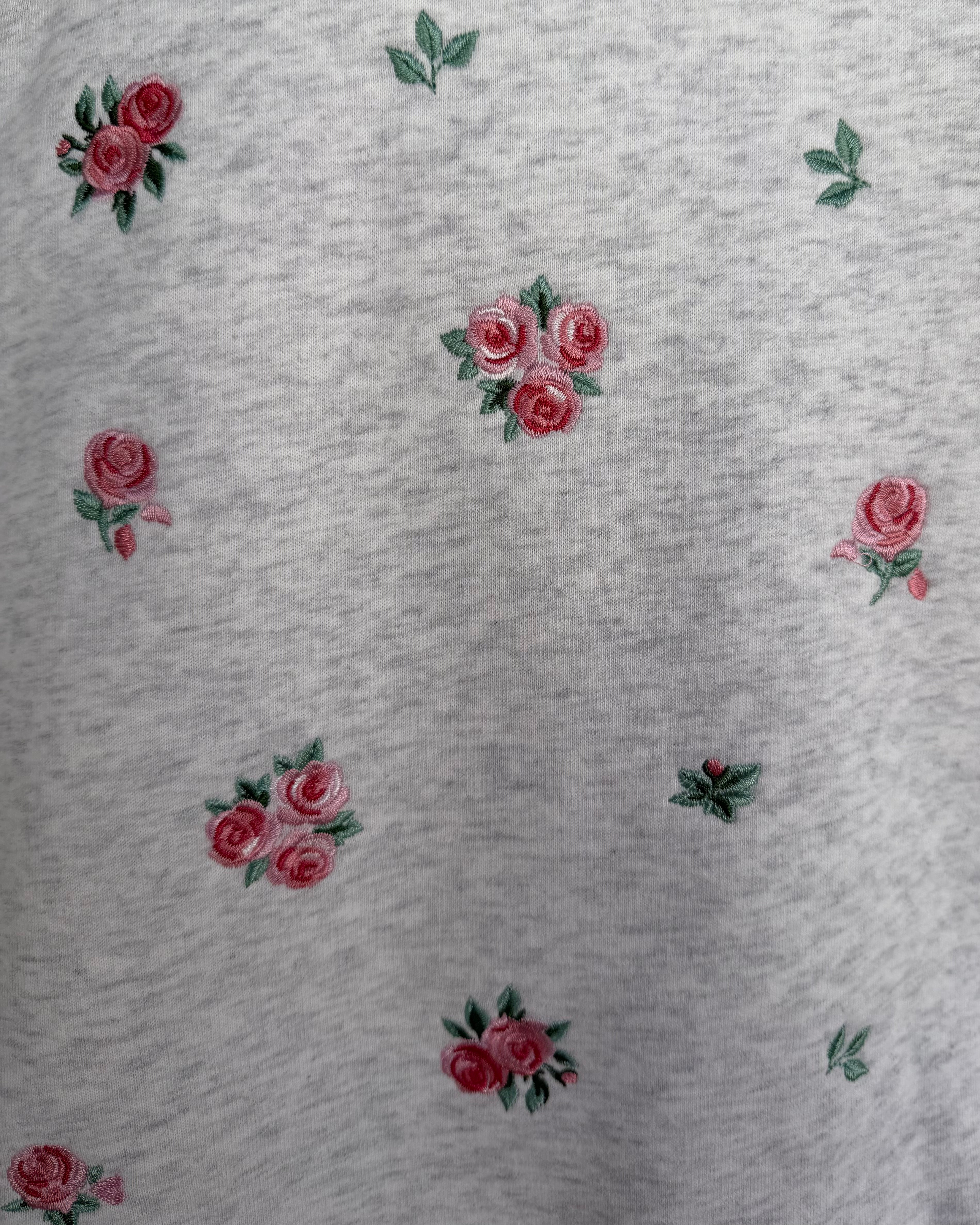 Close-up of a light gray heathered fabric with scattered embroidered pink roses and green leaves.