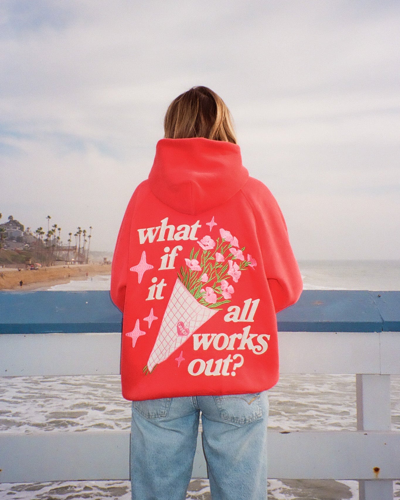 Person wearing a red hoodie with a message and bouquet graphic, standing on a beach.