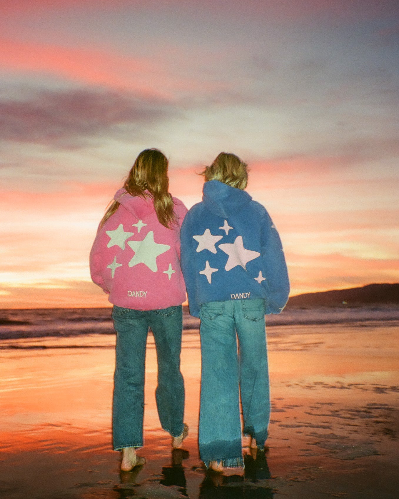 Two people standing on a beach at sunset, wearing colorful hoodies with star designs.