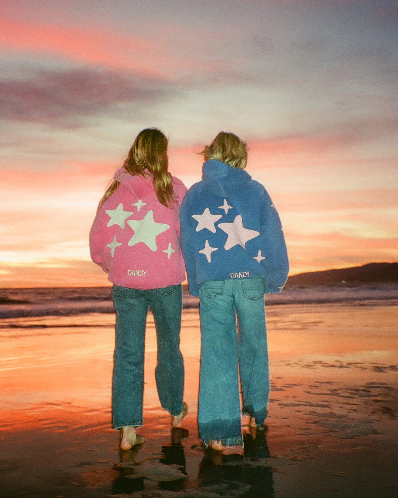 Two people standing on a beach at sunset, wearing colorful hoodies with star designs.