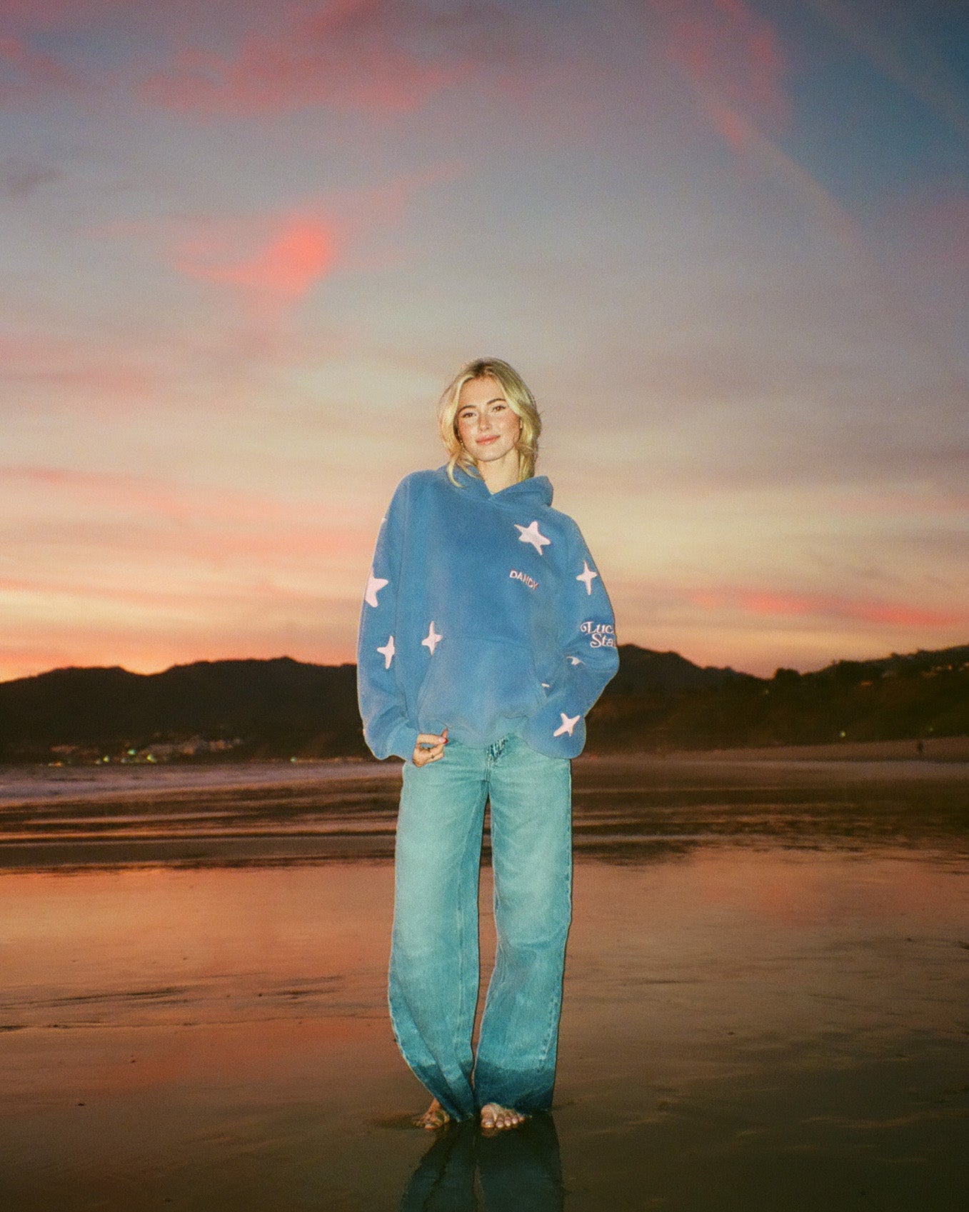 Person wearing a blue sweater and jeans standing on a beach at sunset.