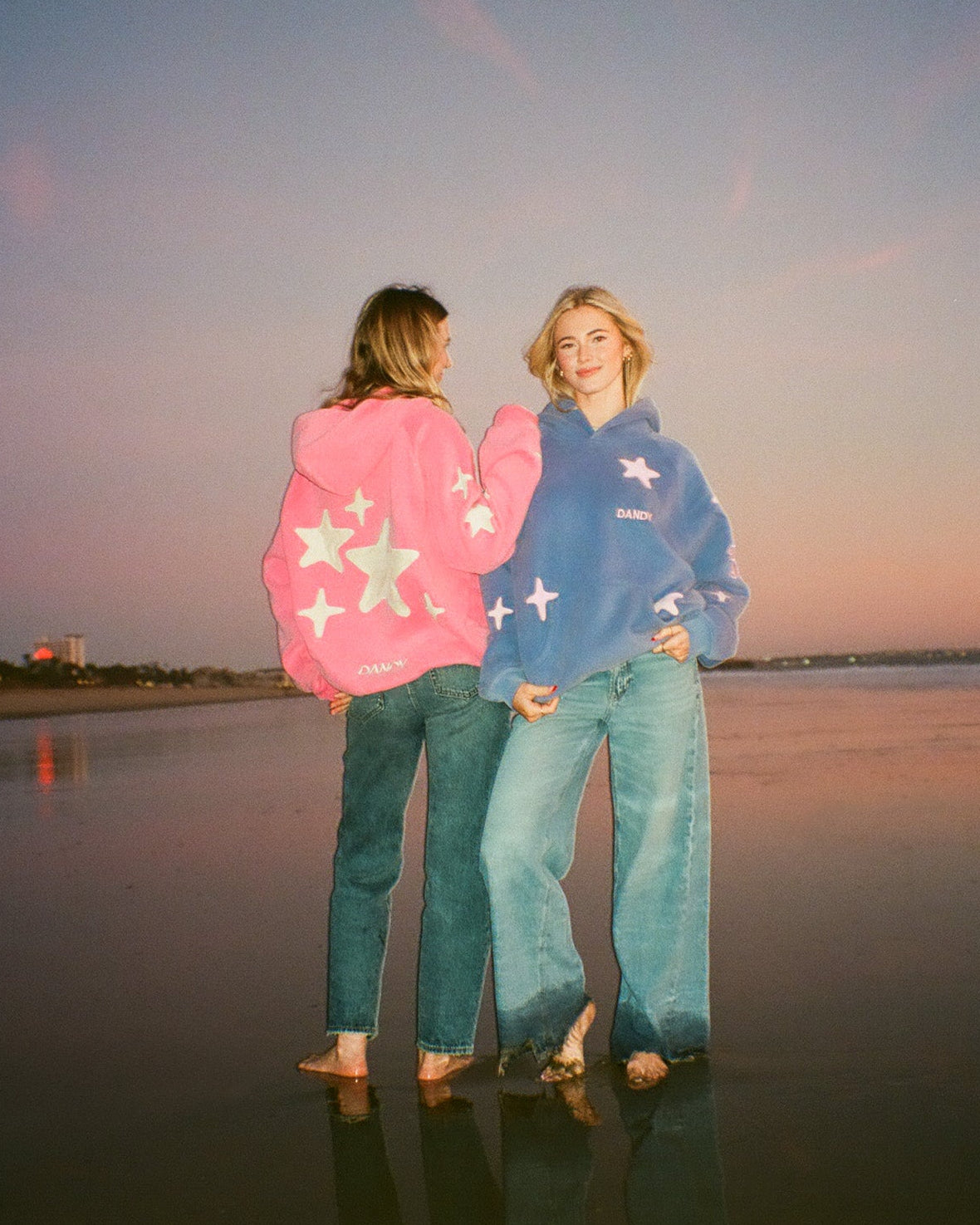 Two people standing by a body of water at sunset, wearing colorful hoodies.