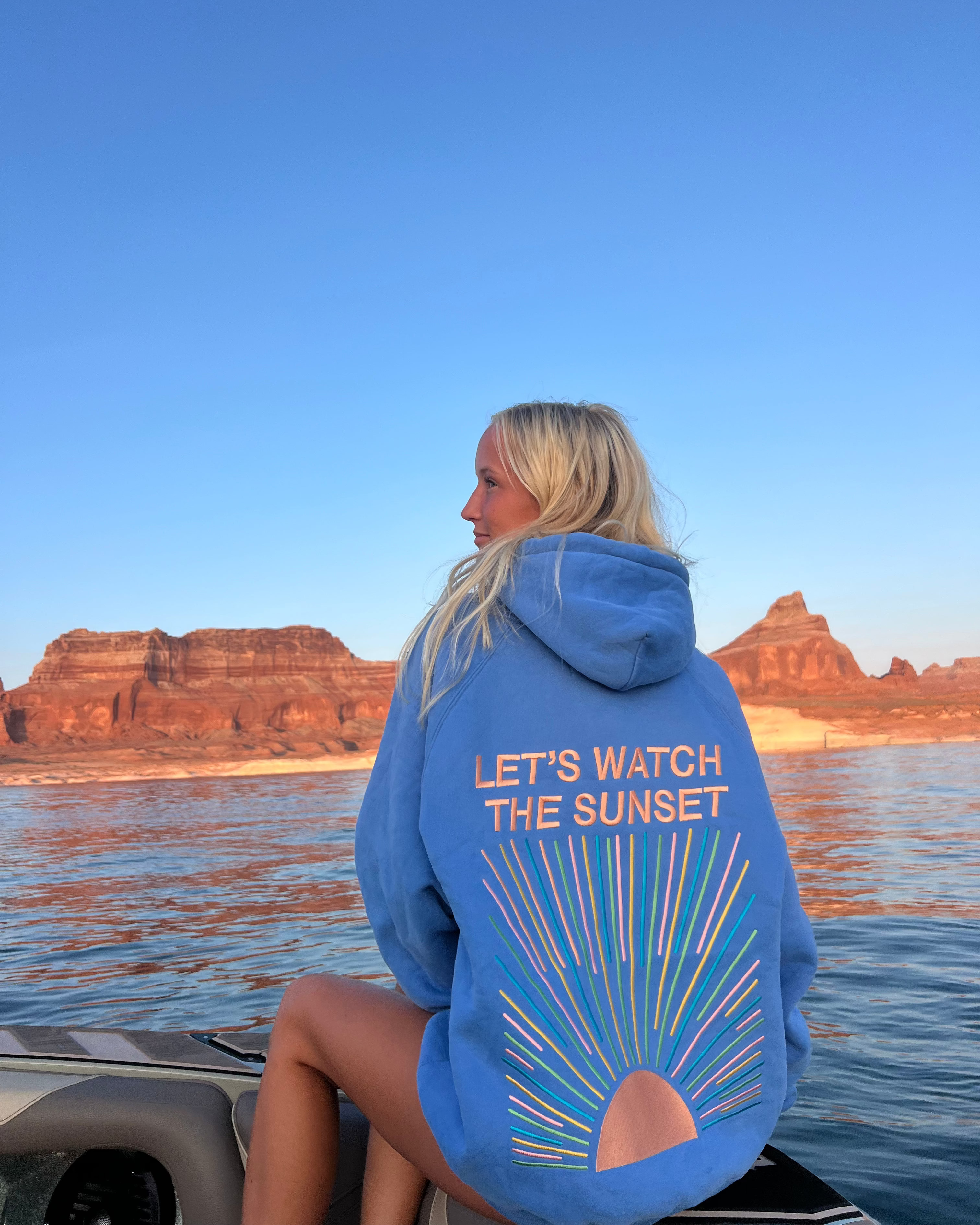 Person wearing a blue hoodie with 'Let's Watch the Sunset' text by a lake with mountains in the background