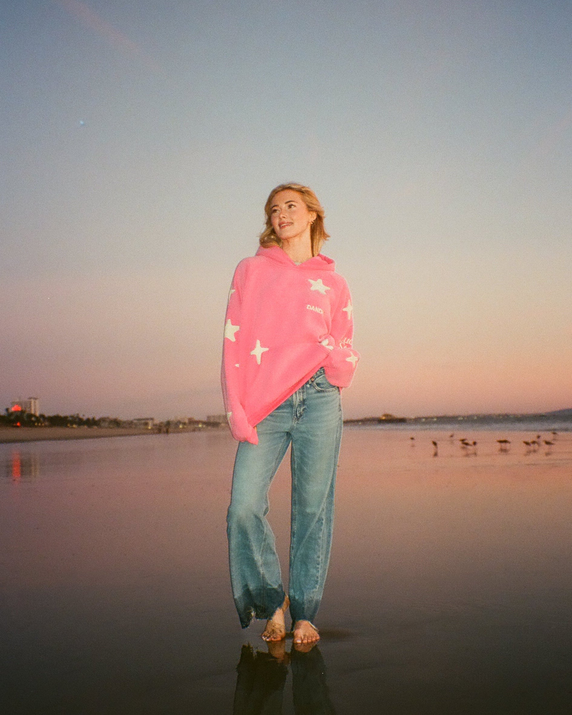 Person standing on a beach at sunset wearing a pink sweater and blue jeans.
