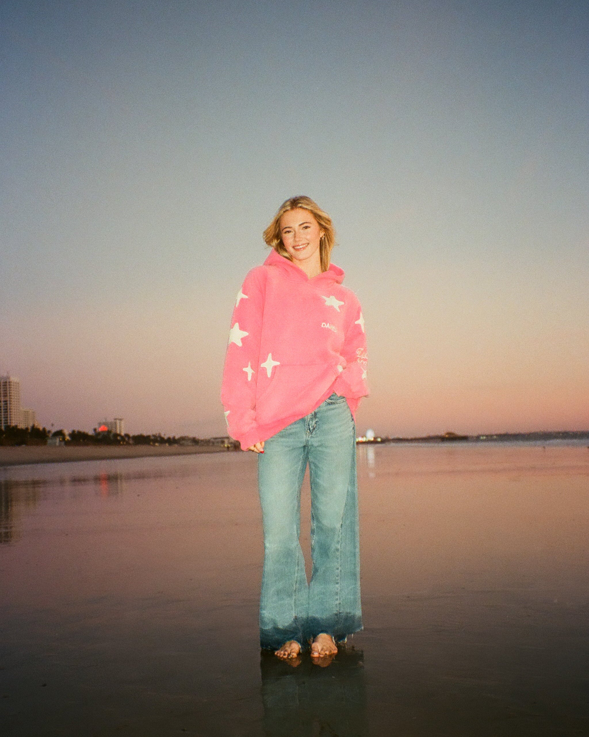 Person wearing a pink sweater and blue jeans standing on a beach at sunset.