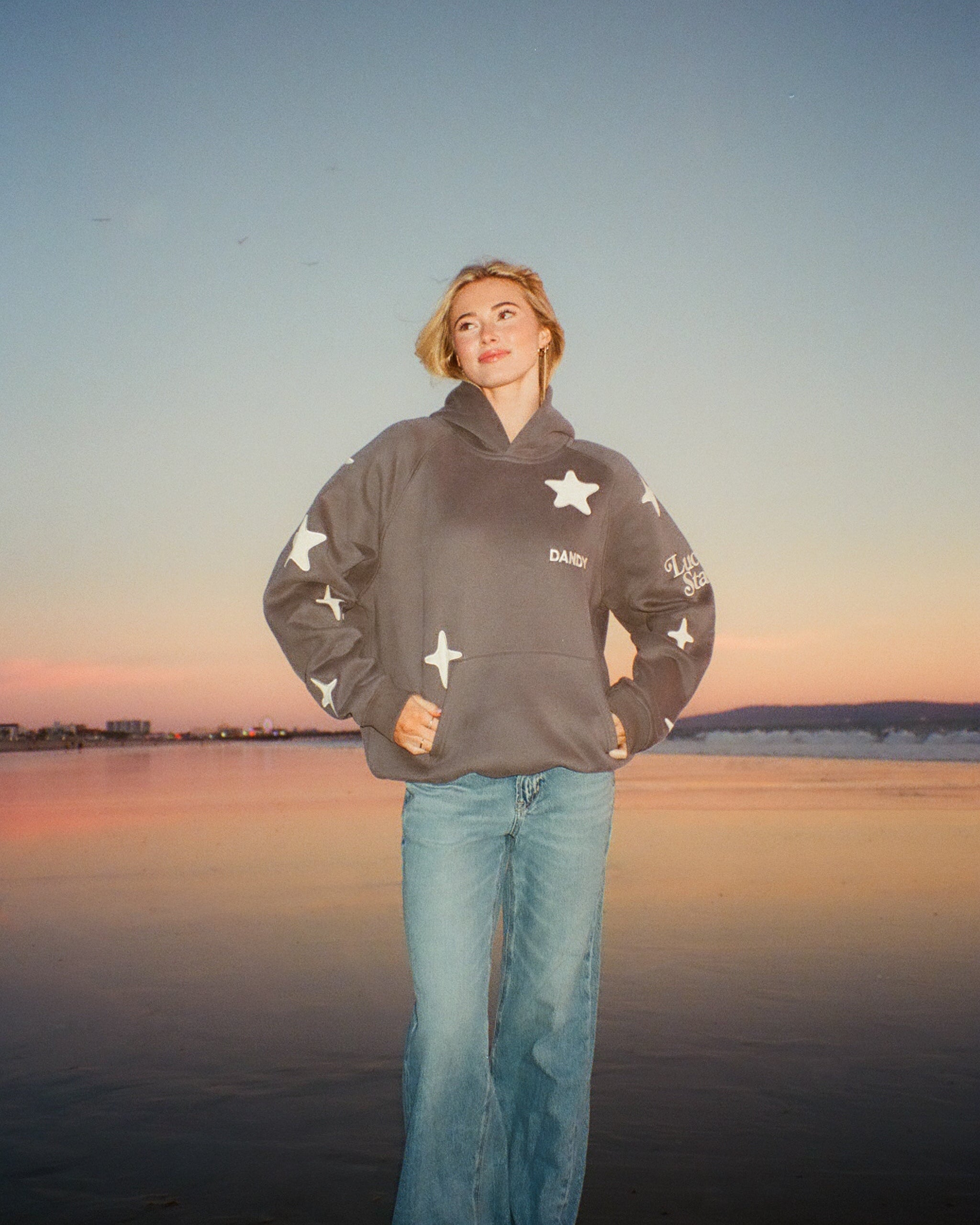 Woman standing on sandy beach at sunset wearing gray star hoodie and jeans