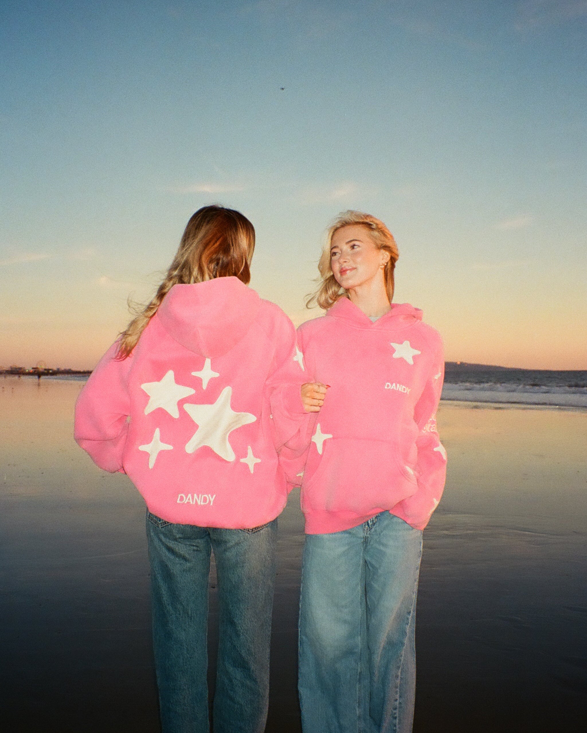 Two people wearing pink hoodies with star patterns on a beach at sunset.