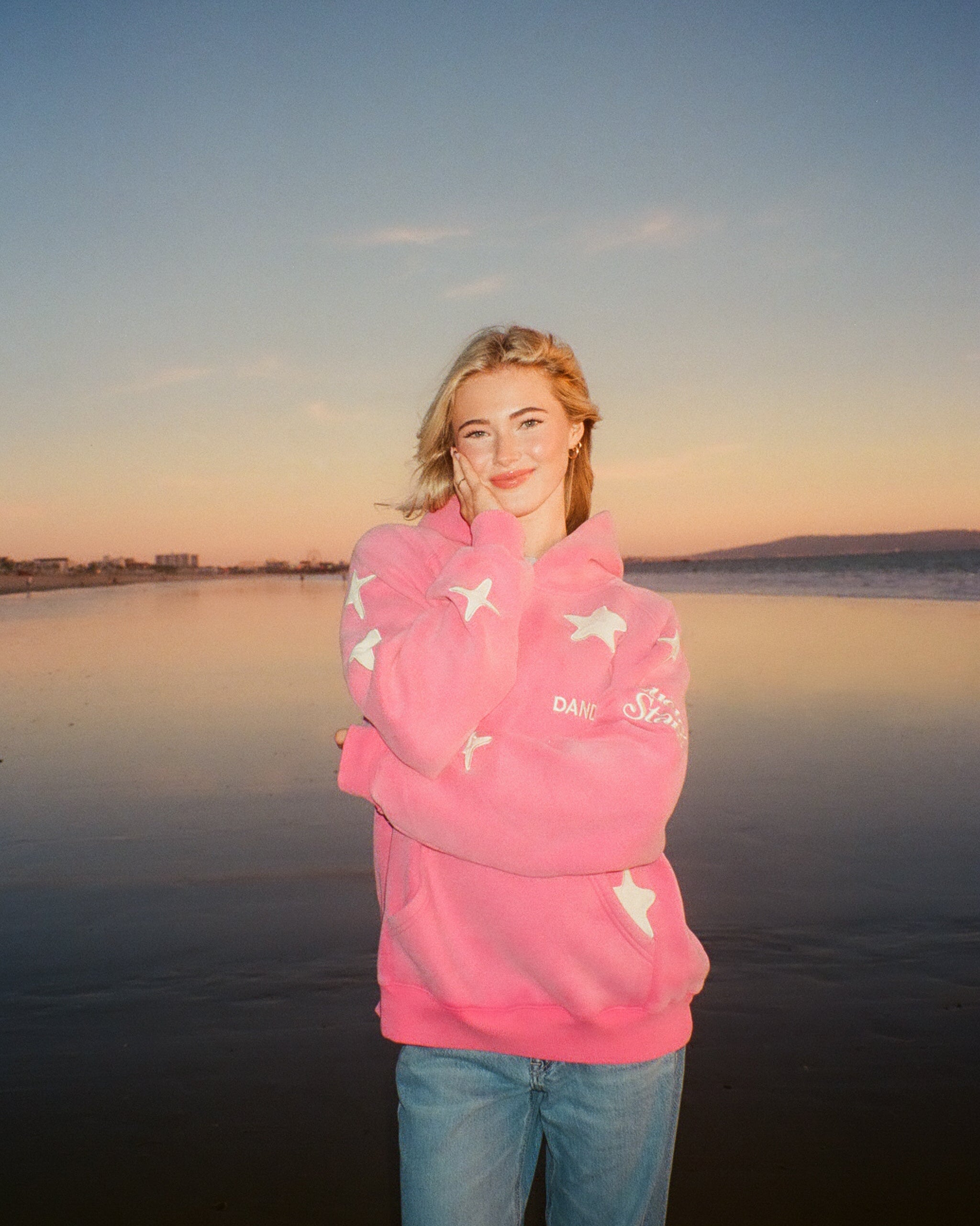 Person wearing a pink hoodie with star patterns and text, standing by a body of water at sunset.