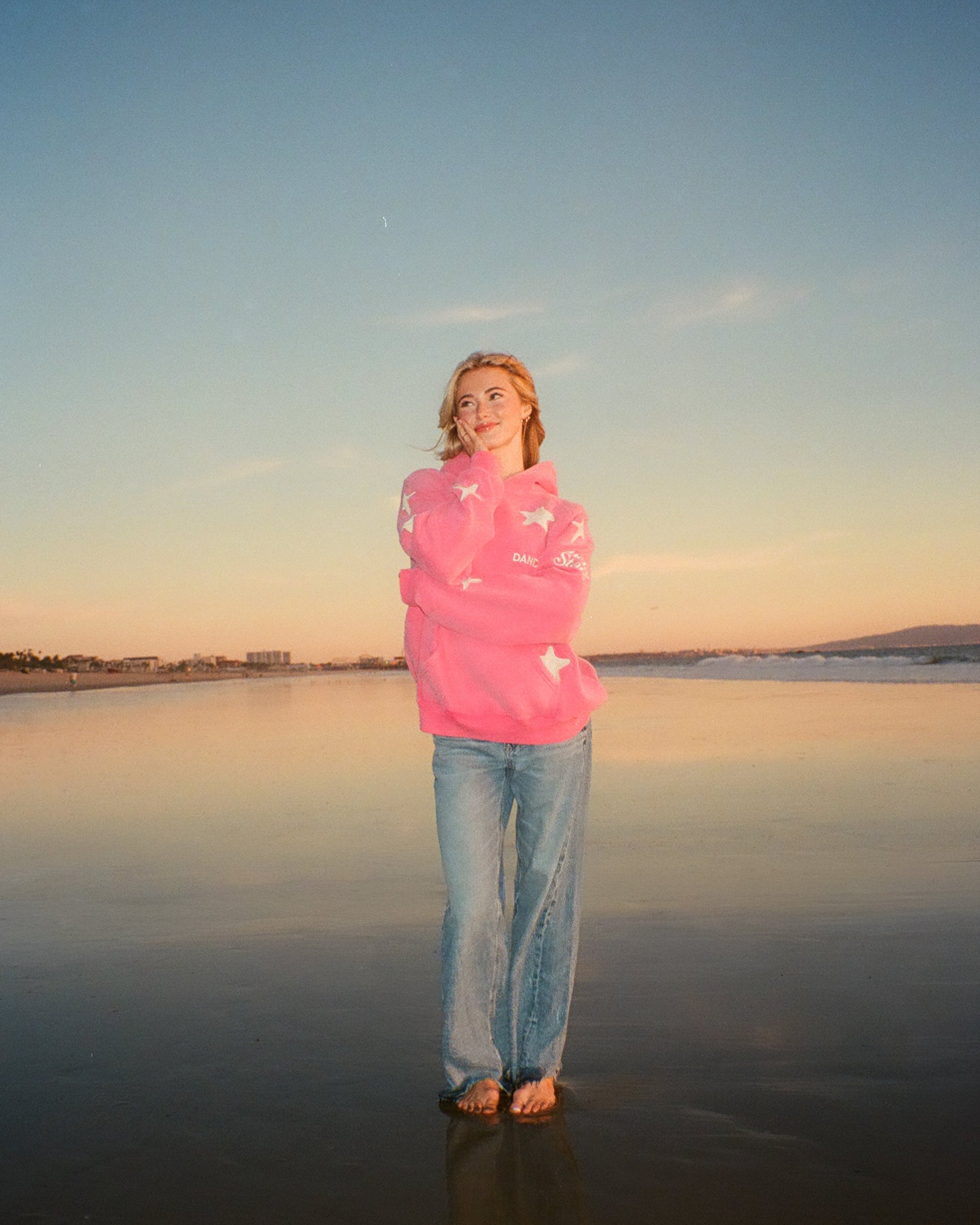 Person standing on a beach at sunset wearing a pink jacket and blue jeans.