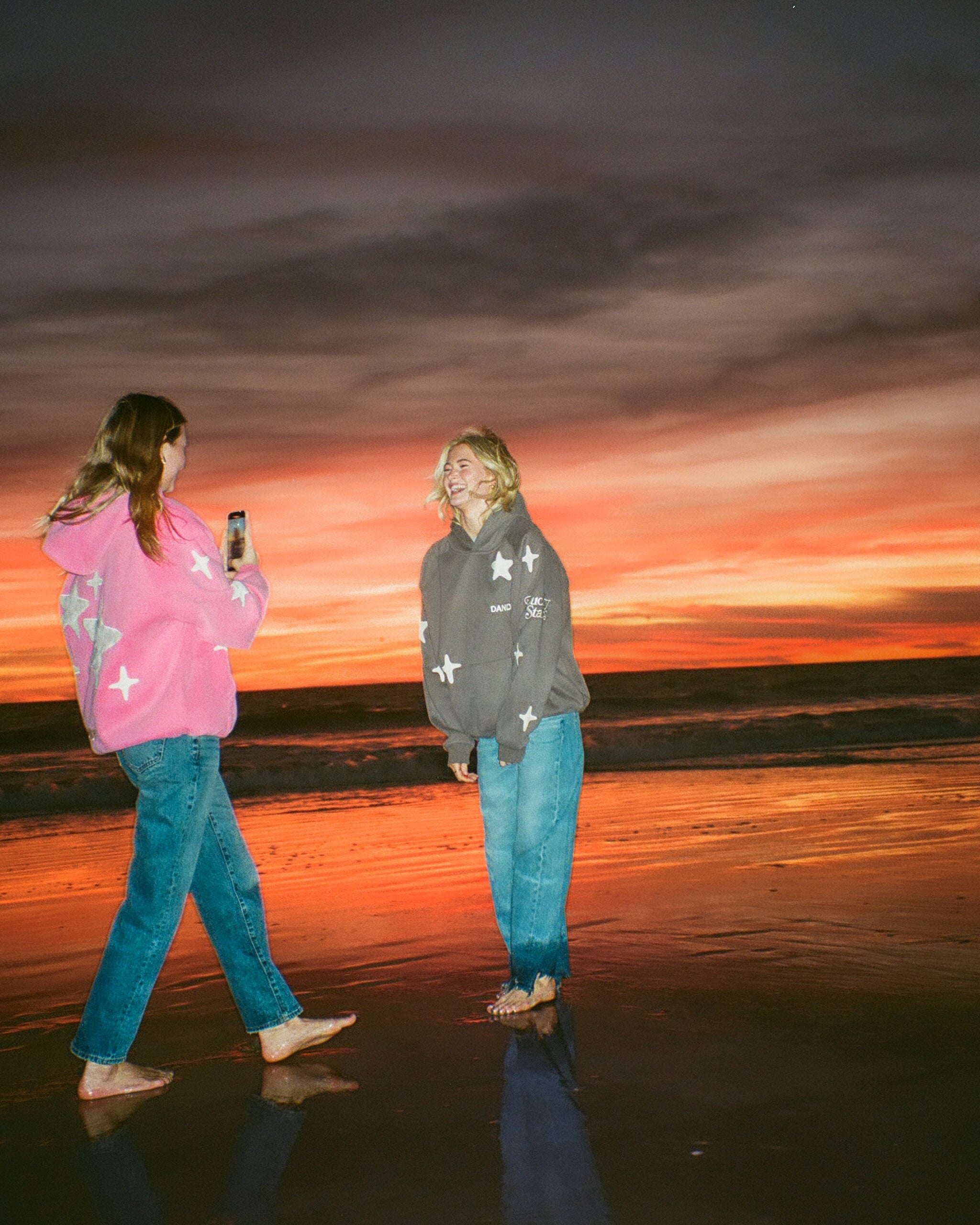 Two girls standing on a beach at sunset wearing star hoodies and jeans