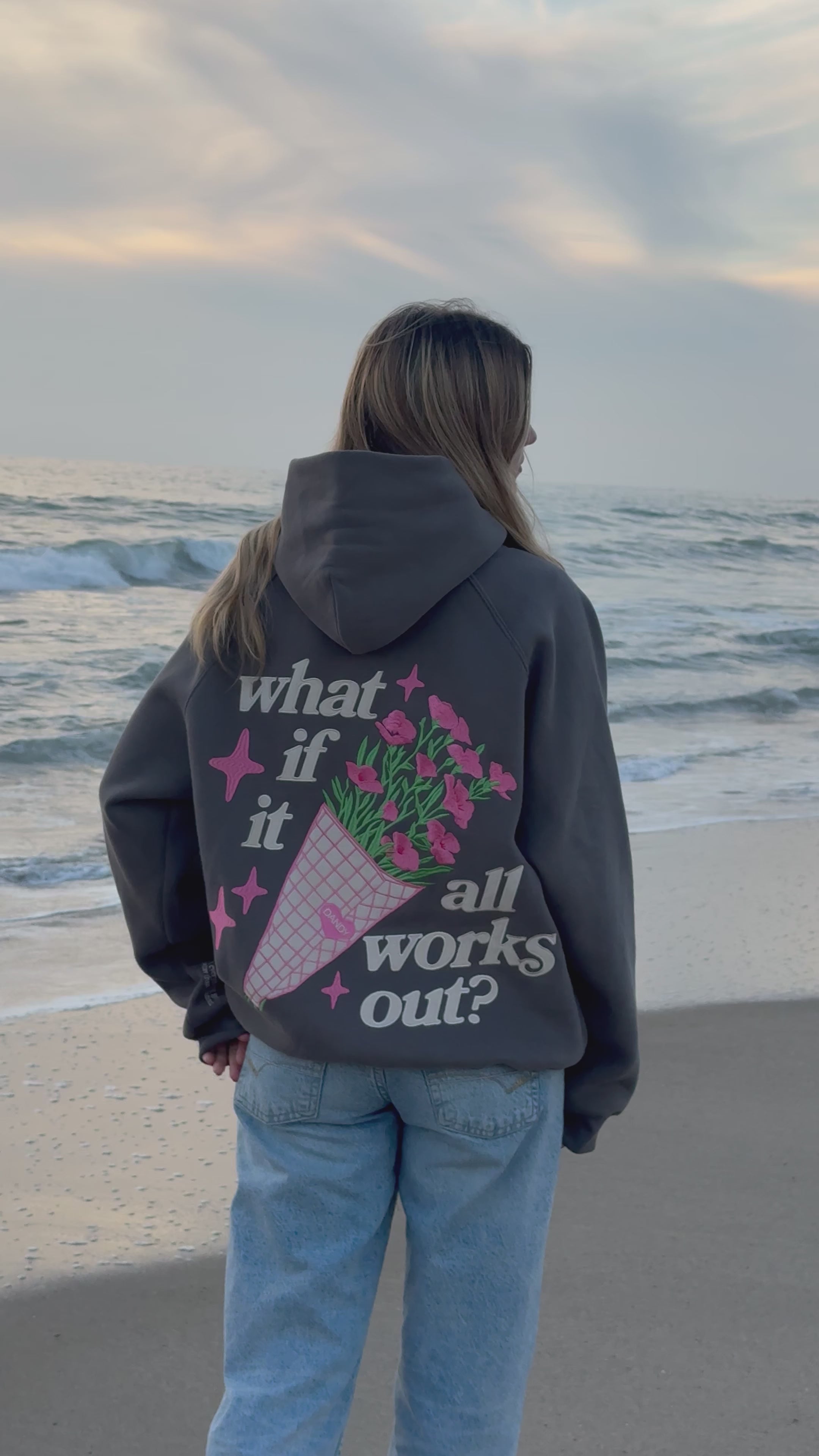 Video of a woman wearing a gray hoodie with flower embroidery on the beach at sunset