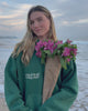 Video showing a woman walking on a sandy beach holding flowers and wearing a green hoodie